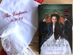 The Empress by Gigi Griffis alongside a handkerchief embroidered with the book title and date